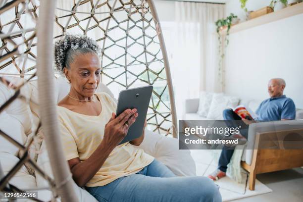 pareja mayor en sala de estar, hombre leyendo libro y mujer usando tableta digital - old man afro fotografías e imágenes de stock