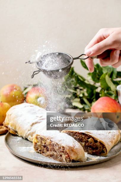 Homemade sliced classic apple strudel sprinkling by icing sugar from sieve in hand. On ceramic plate with fresh apples. Green leaves and cinnamon...