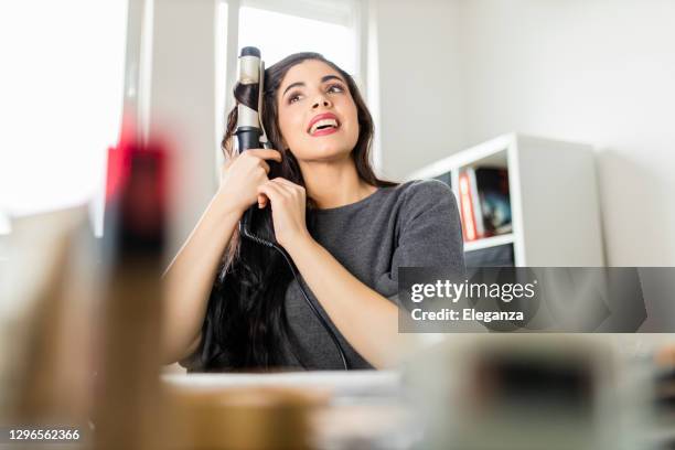 frau schaut auf computer und mit ihrem lockenbügeleisen, während sie am schminktisch sitzt. frau macht ihre haare, indem sie ein tutorial im internet zum zeitpunkt der corona-virus-pandemie. - lockenstab stock-fotos und bilder
