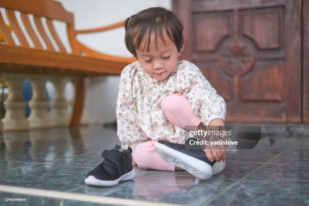 Cute little Asian toddler girl sitting and concentrate on putting on her black shoes, sneakers, Get toddler out the door, Encourage Self-Help Skills in Children concept