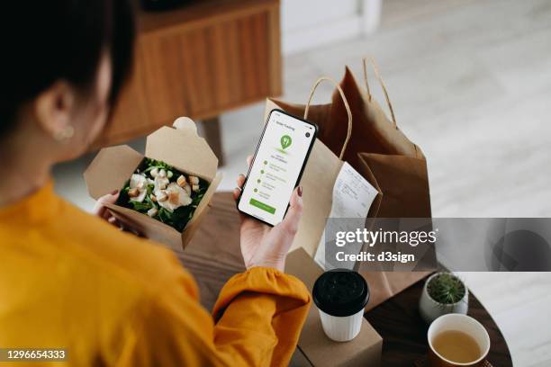 over the shoulder view of young asian woman ordering home delivery takeaway food with mobile app on smartphone, tracking status and confirming the food order. enjoying a box of fresh grilled chicken vegetable salad. technology makes life so much easier - food delivery fotografías e imágenes de stock