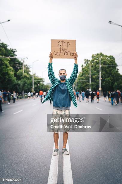 young man with protective face mask, surrounded by a crowd, holding a poster in his hands. young activist / protester against the corruption. human rights and social issues concept. - anti graft stock pictures, royalty-free photos & images