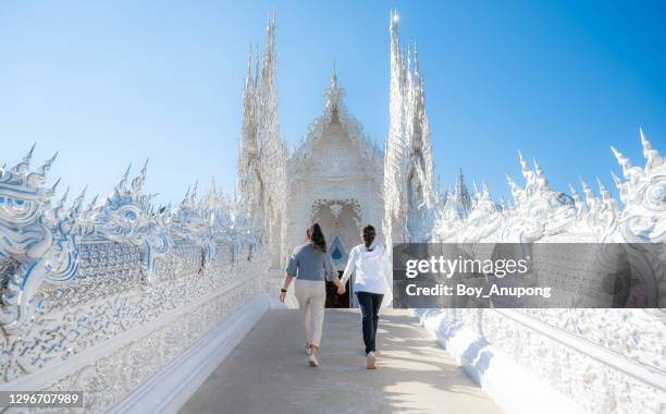 asian tourist women holding other hand and walking together to the main building of wat rong khun (white temple) an iconic tourist attraction in chiang rai province of thailand. - wat rong khun stock pictures, royalty-free photos & images