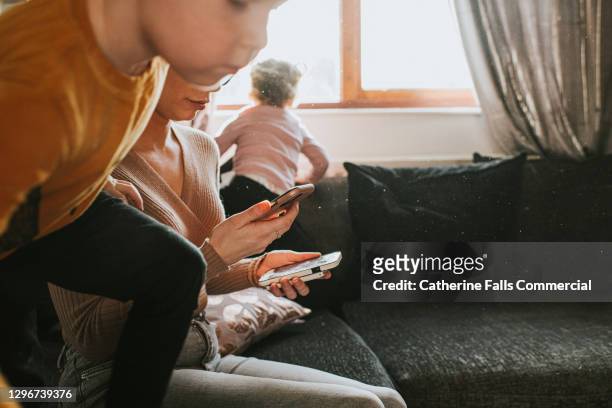 a mum holds two mobile phone and attempts to work from home as kids jump around her - welfare stock pictures, royalty-free photos & images