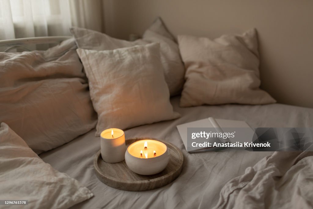 Scented candles in ceramic bowls on linen bed with book at home.