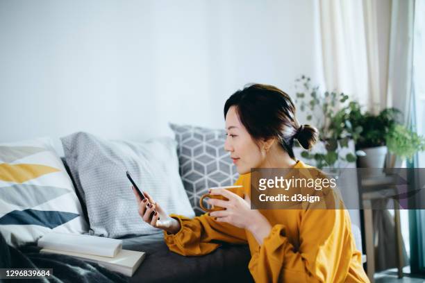 beautiful smiling young asian woman chilling at cozy home, sitting on the floor by the sofa, enjoying a cup of coffee and using smartphone - dating stockfoto's en -beelden