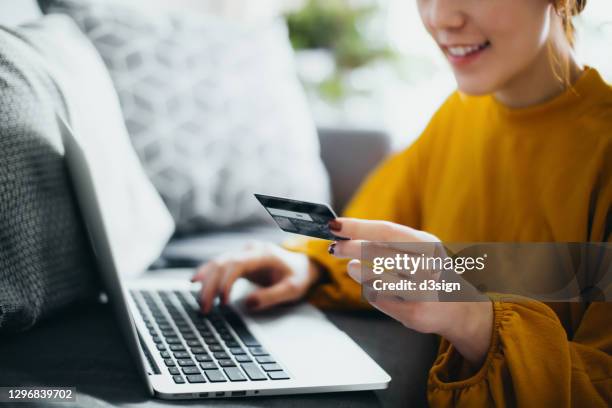 close up of beautiful smiling young asian woman sitting on the floor by the sofa, managing financial bill payment with laptop and credit card on hand at cozy home. technology makes life so much easier - credit card stock-fotos und bilder