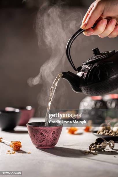 hand pouring cup of green tea from black cast iron teapot into pink cup. lot of steam - herbal tea bag stock pictures, royalty-free photos & images