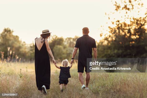 parent and baby girl in black casual clothes walking in summer park holding hands on sunset. view from the back. family day. - girls hands behind back stock-fotos und bilder