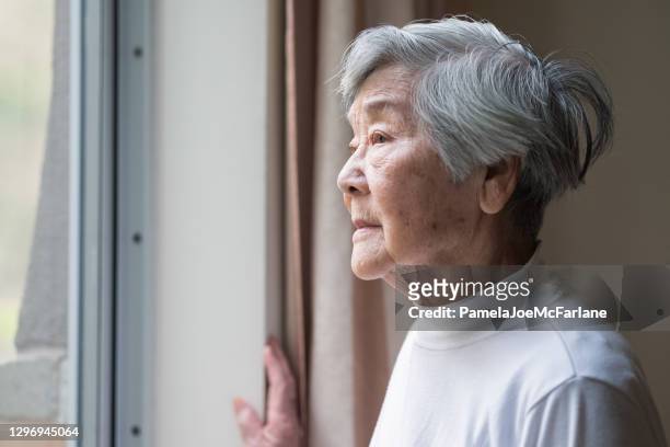 grave donna anziana asiatica negli anni '90 guardando fuori dalla finestra - solitudine foto e immagini stock