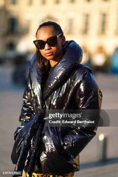 Emilie Joseph wears sunglasses, a black shiny oversized winter puffer jacket with oversized lapels from Gina Tricot, yellow and black floral print...