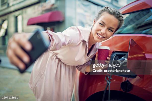 la joven está tomando selfie / haciendo videollamada hasta cargar su coche eléctrico - red car wire fotografías e imágenes de stock