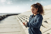 Young purposeful curly-haired fitness woman in sportswear looking at watch on city beach