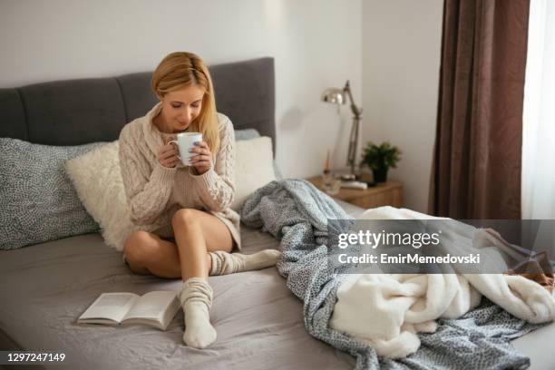 young woman enjoying a good book at home while drinking coffee in bed - cosy bed stock pictures, royalty-free photos & images
