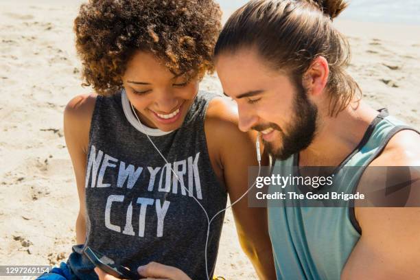 friends looking at smart phone while sitting on sand at beach - beach music stock pictures, royalty-free photos & images