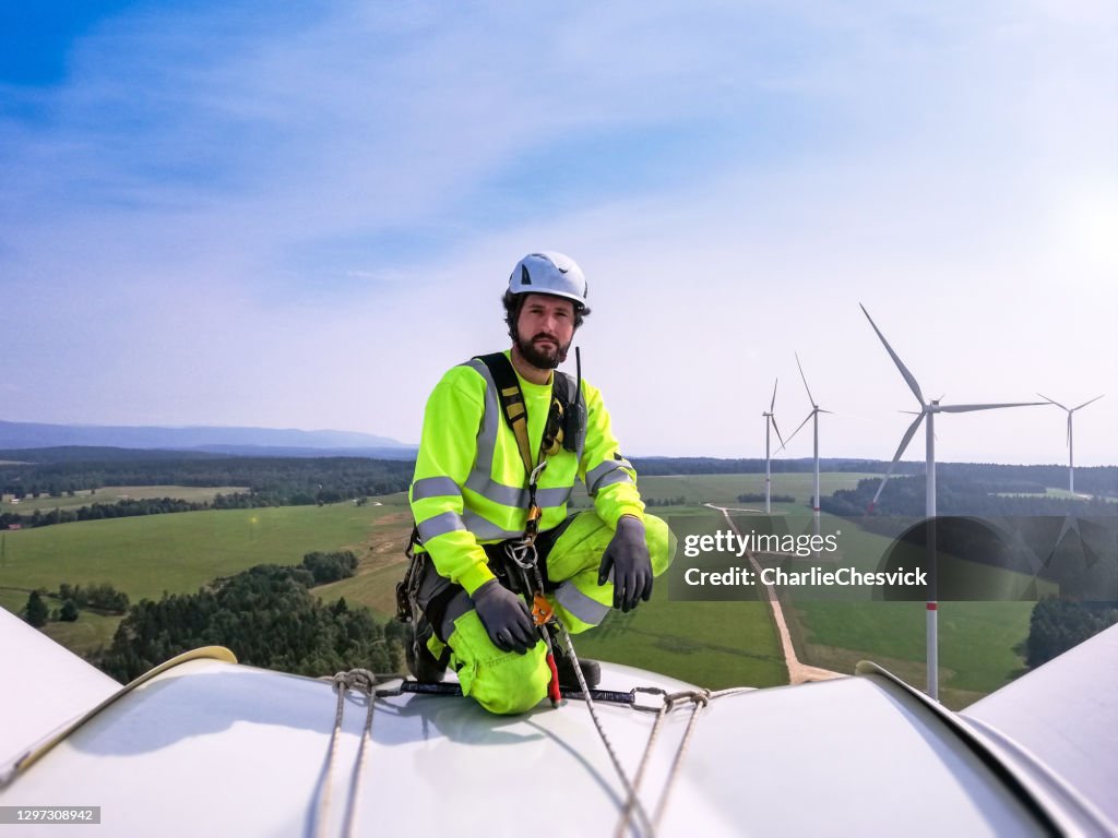 Industriële klimmer, de technicus die van de kabeltoegang op enorme onshore windturbine knielt terwijl het voorbereiden voor bladinspectie met windpark achter hem