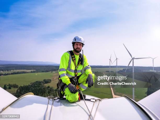 industrial climber, rope access technician kneeling on enormous onshore wind-turbine while preparing for blade inspection with wind farm behind him - turbina eólica imagens e fotografias de stock