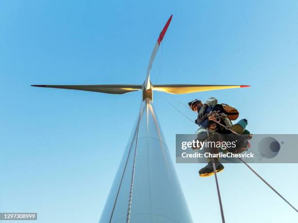 vista de ángulo bajo en el técnico de acceso a cuerdas, escalador industrial colgando en la cuerda del aerogenerador iluminado por la puesta de sol y mirando a la cámara con mangas de zapatos - turbina fotografías e imágenes de stock