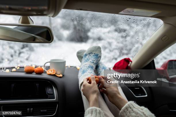 woman's feet in warm woolen socks with tangerines, christmas garlands and coffee cup on car dashboard over snow view. having weekend trip in winter forest. freedom travel concept. - feet christmas stock-fotos und bilder