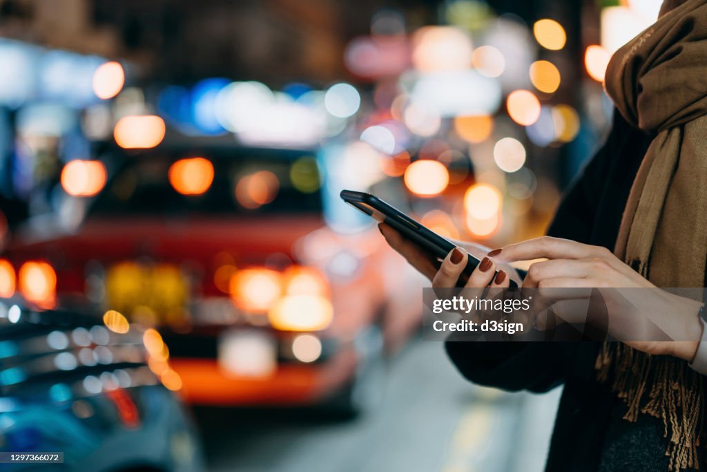 Close up of young woman using mobile app device on smartphone to arrange taxi ride in downtown city street, with illuminated busy city traffic scene during rush hour with traffic congestion in the evening