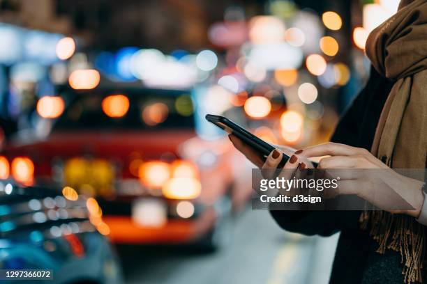 close up of young woman using mobile app device on smartphone to arrange taxi ride in downtown city street, with illuminated busy city traffic scene during rush hour with traffic congestion in the evening - taxi stock pictures, royalty-free photos & images