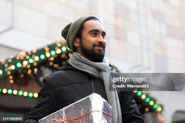 smiling young man holding christmas present while standing against building in city - kühlbox stock-fotos und bilder