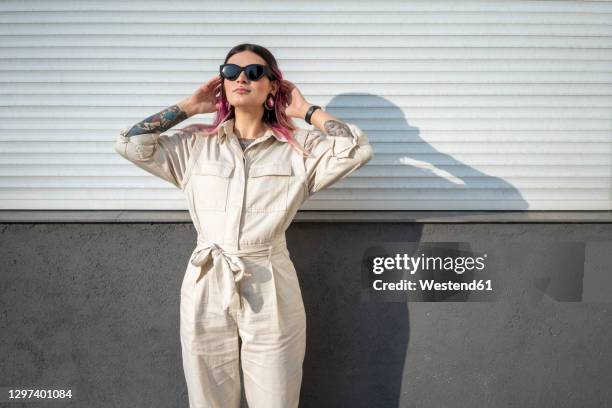fashionable woman with hand in hair against wall on sunny day - jumpsuit stockfoto's en -beelden