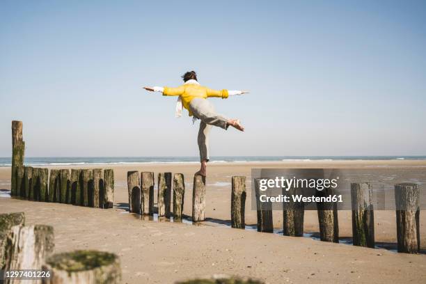 woman with arms outstretched balancing on wooden posts at beach against clear sky - standing on one leg fotografías e imágenes de stock