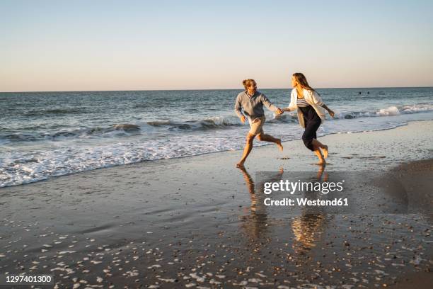 happy couple holding hands while running at beach against clear sky during sunset - running netherlands stockfoto's en -beelden