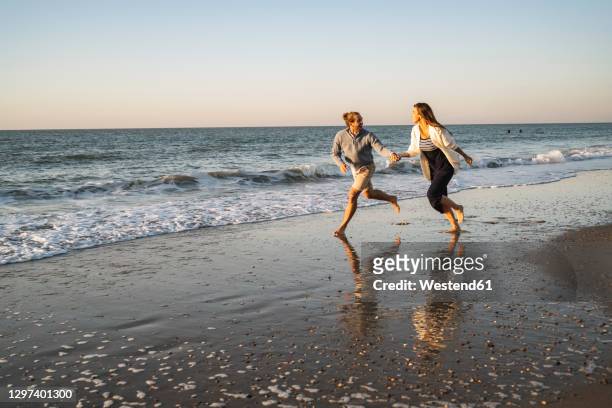 happy couple holding hands while running at beach against clear sky during sunset - running netherlands stock pictures, royalty-free photos & images