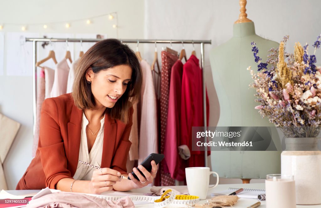 Female fashion designer using smart phone while leaning on desk in atelier