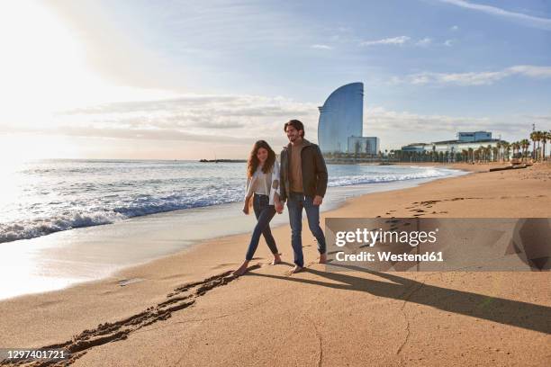 girlfriend and boyfriend holding hand while walking on sand at beach - barcelona coast stock pictures, royalty-free photos & images