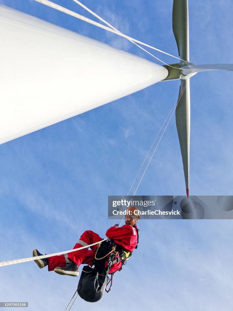 View from bottom wind-turbine and industrial climber rope access technicians abseiling down from blade on the ropes, looking down, dramatic sky and sunny day.