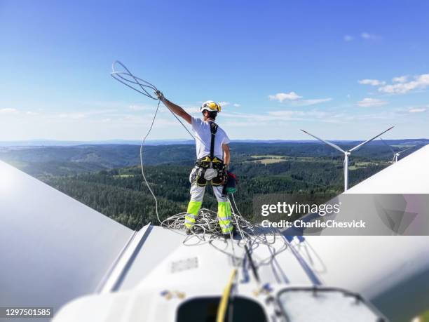 vista posteriore sul tecnico professionista di accesso alle funi in piedi sul tetto (mozzo) della turbina eolica e tirando su la corda. il sole è dietro la turbina eolica. - energia eolica foto e immagini stock