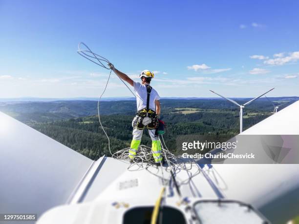 rear view on professional rope access technician standing on roof (hub) of wind turbine and pulling rope up. sun is behind wind turbine. - image technique imagens e fotografias de stock