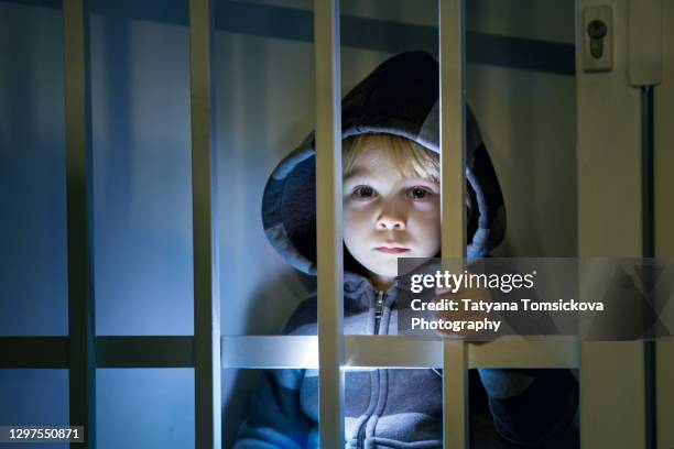 small toddler child, blond boy, standing locked behind bars, child punishment, sad kid - child behind bars stock-fotos und bilder