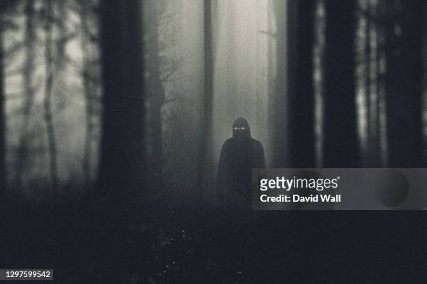 a grainy horror concept. of a dark hooded figure with glowing eyes standing in a forest. with an abstract blurred edit. - spooky stock-fotos und bilder
