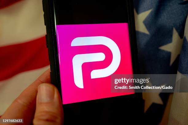 Illustrative image of human hand with mobile device showing logo for the social media platform Parler with American flag visible in background,...