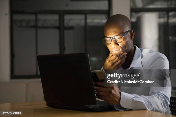 reife schwarze mann bei der arbeit an seinem laptop und handy in der nacht - fake news stock-fotos und bilder