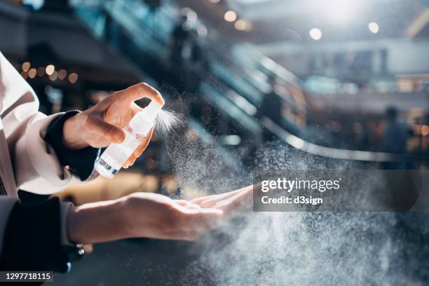 close up of young asian woman sanitizing her hand with disinfectant spray while shopping in a shopping mall against sunlight, with escalator and shoppers in background - hygiene stock-fotos und bilder