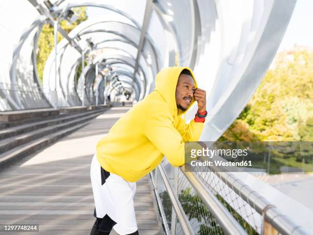 thoughtful businessman wearing yellow hooded shirt leaning on walkway railing - forward athlete stockfoto's en -beelden