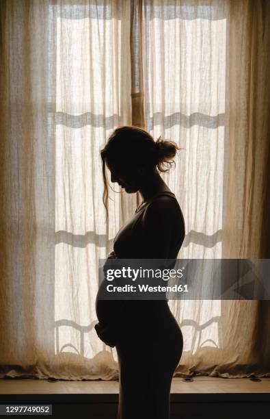 pregnant woman touching belly while standing by window at home - gravidanza foto e immagini stock