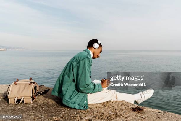 young man with headphones writing in diary by sea and sky - man in denim jacket stock pictures, royalty-free photos & images