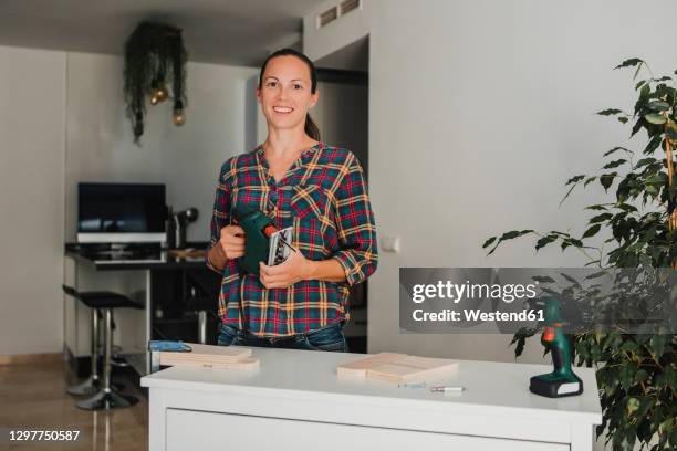 mid adult woman holding electric jigsaw while standing by table at home - serra tico tico serra elétrica - fotografias e filmes do acervo