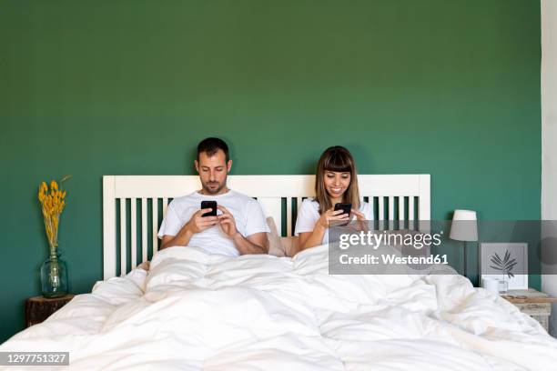 couple using their mobile phones while sitting in bed at home - couple on bed stock-fotos und bilder