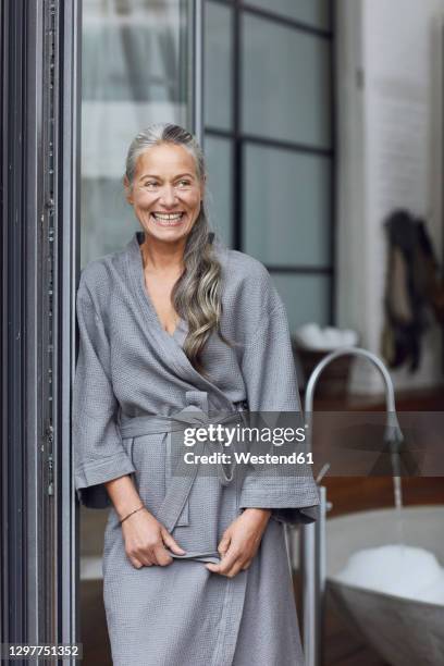 smiling mature woman wearing bathrobe leaning by glass door in bathroom - bathrobe stockfoto's en -beelden