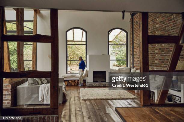 mature woman with hands in pockets looking through window at home - holzbalken stock-fotos und bilder