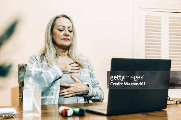 senior woman doing breathing exercise during online consultation at home - adem inhouden stockfoto's en -beelden