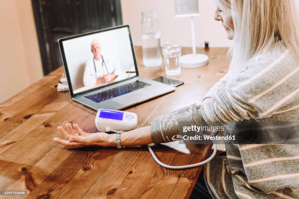Senior woman checking own blood pressure under guidance of doctor on video call at home