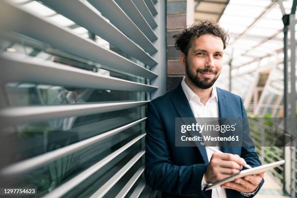 smiling businessman with digital tablet standing by wall in office - digitized pen stock-fotos und bilder